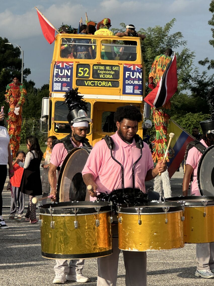 Musicians play drums in a lively street parade with a decorated double-decker bus in the background.